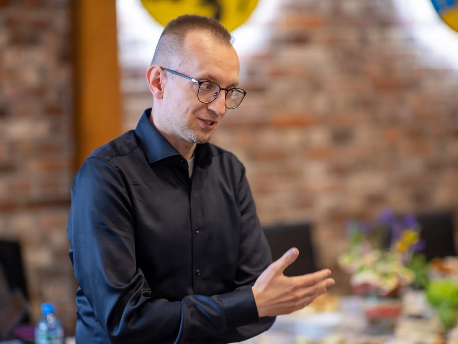 Osoba z zespołu Fundacji w ciemnej koszuli opowiada coś słuchaczom podczas spotkania w sali z ceglaną ścianą